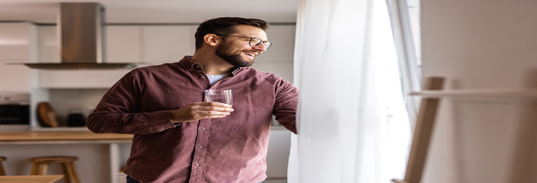 A smiling man stands in a bright, modern kitchen, holding a glass of water while looking out a sunlit window, creating a calm and welcoming morning atmosphere.